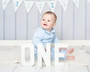 A little boy dressed in blue poses behind some wooden letters that spell ONE in front of a blue background at a professional first birthday photo shoot taking place at my East Grinstead studio