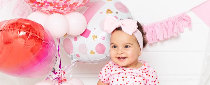 A little girl sits in a dress with hearts on it in front of a bunch of balloons with a cake in front of her at her first birthday photo shoot in West Sussex