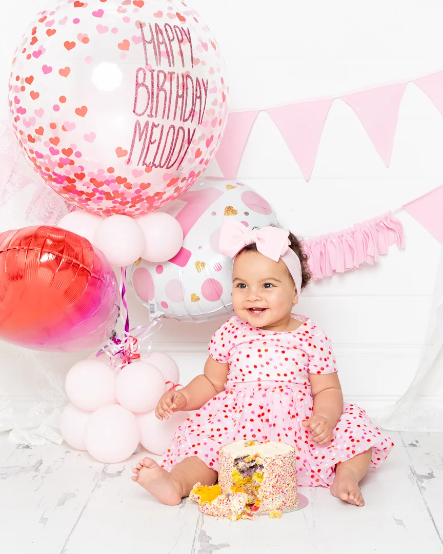 A little girl sits in a dress with hearts on it in front of a bunch of balloons with a cake in front of her at her first birthday photo shoot in West Sussex