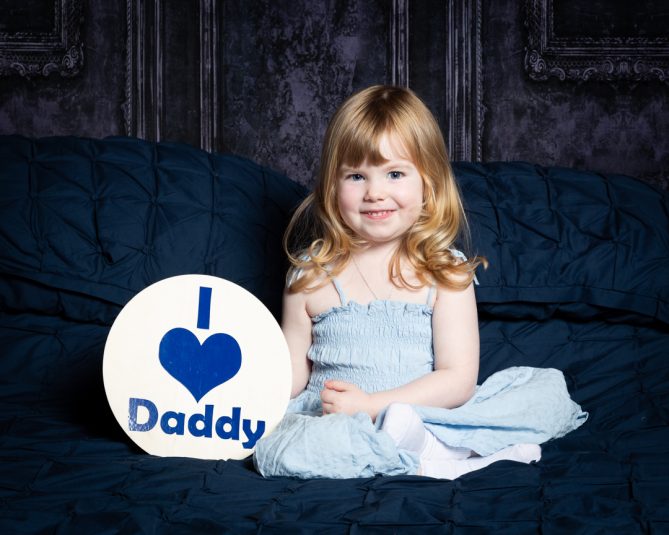 a little girl poses smiling on a bed with a sign saying I love daddy at a father's Day photo shoot