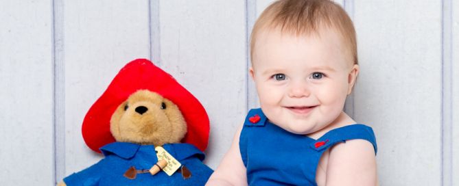 A baby dressed in a blue outfit sits with a Paddington toy holding a miniature suitcase