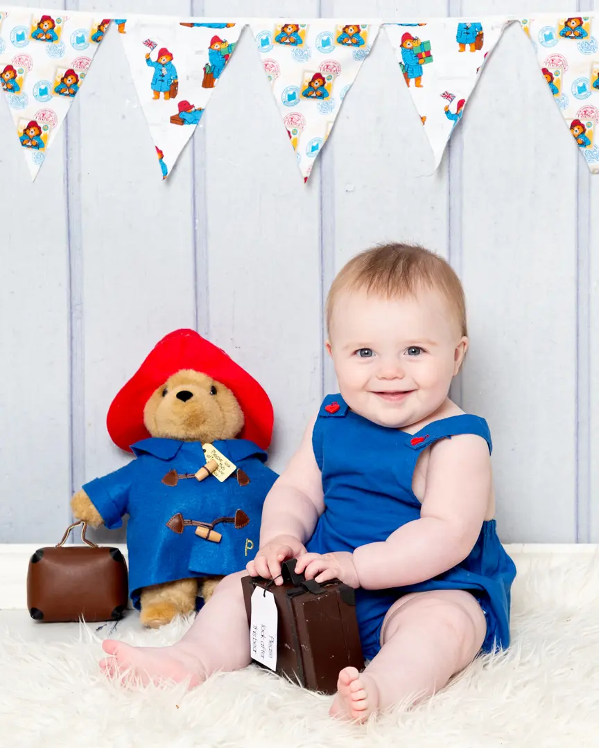 A baby dressed in a blue outfit sits with a Paddington toy holding a miniature suitcase