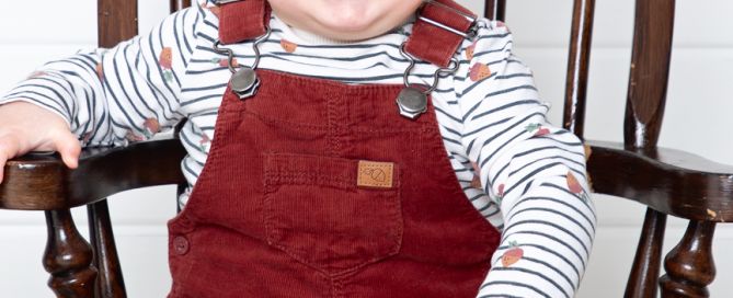 a little boy in dungarees sits in a miniature wooden chair