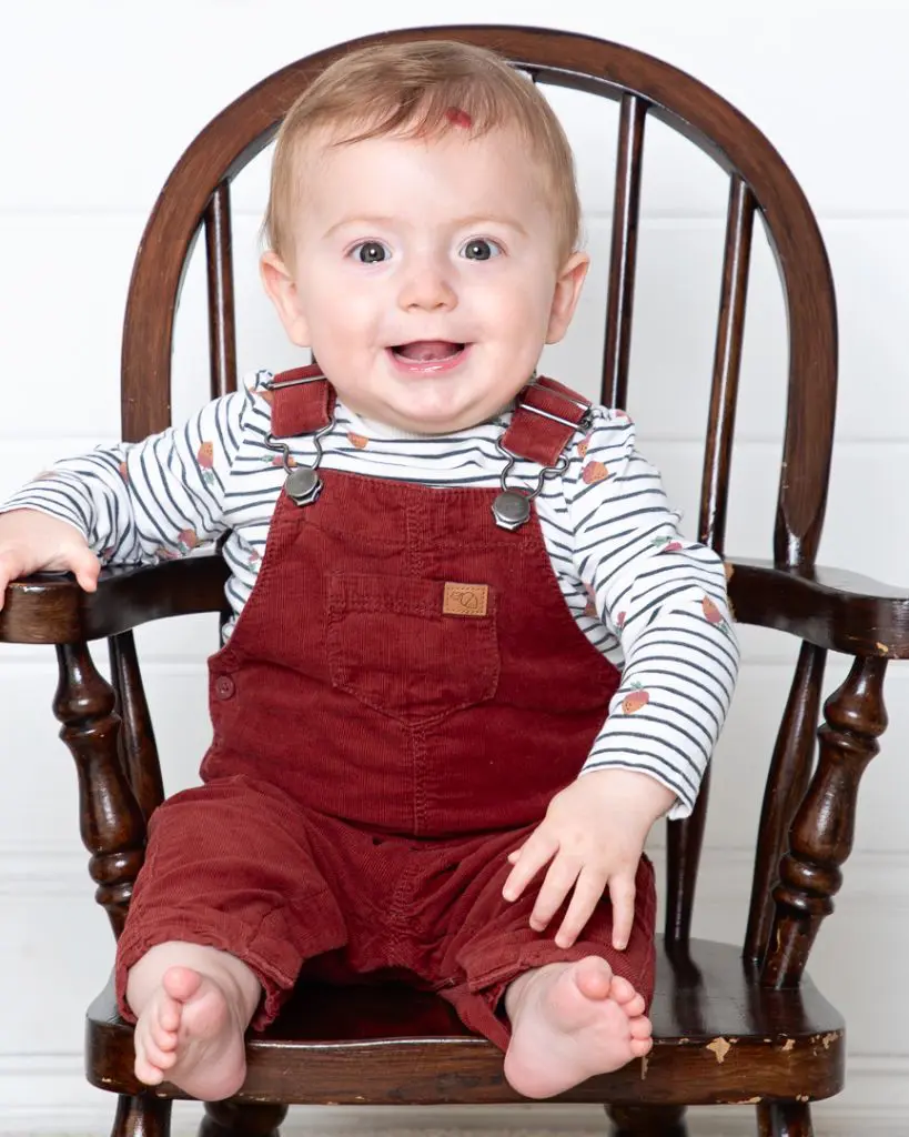 a little boy in dungarees sits in a miniature wooden chair