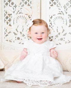 A baby girl is sitting in front of a Boho style backdrop wearing a white dress at a professional baby photo shoot