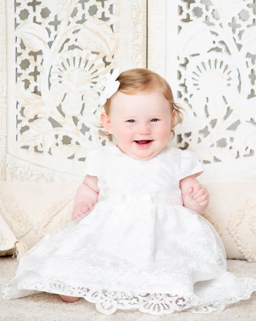 Baby Photography West Sussex a baby in a white dress against a boho screen