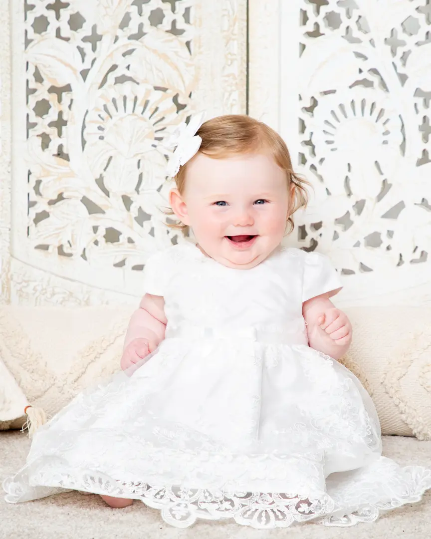 a baby in a white dress against a boho screen