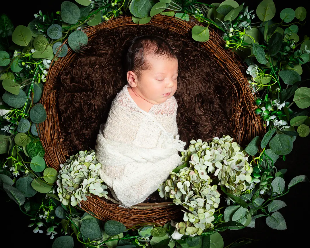 a newborn swaddled in white sleeping in a nest prop surrounded by greenery