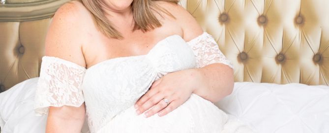 a pregnant woman sits on a bed in a white dress