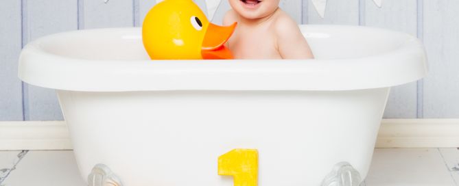 a little boy sits in a white rolltop bath holding a giant yellow duck at a first birthday photo shoot