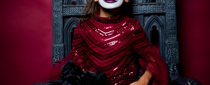 A girl in a gothic red dress with spooky face paint sits on a sjkull throne at a professional photo shoot