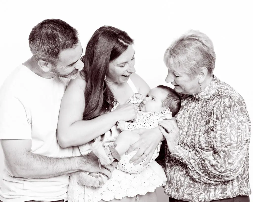 Caprturing Family Moments in East Grinstead A family of two parents and a grandparent look down at a small baby as they hold her in their arms - a black and white image taken in my East Grinstead portrait studio.