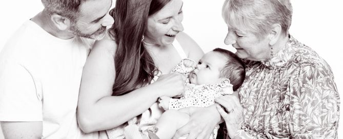 A family of two parents and a grandparent look down at a small baby as they hold her in their arms - a black and white image taken in my East Grinstead portrait studio.
