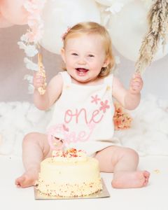  A little girl is holding her arm in the air as she has fun playing with her cake at a first birthday photo shoot