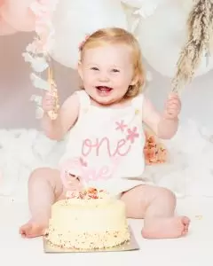  A little girl is holding her arm in the air as she has fun playing with her cake at a first birthday photo shoot