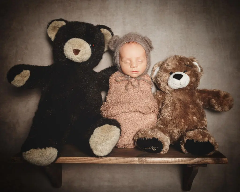A newborn dressed as a teddy bear sitting on a shelf with two actual teddy bears