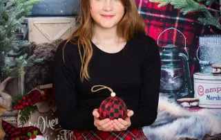 a young girl holds a christmas bauble as she poses for a plaid themed christmas photo