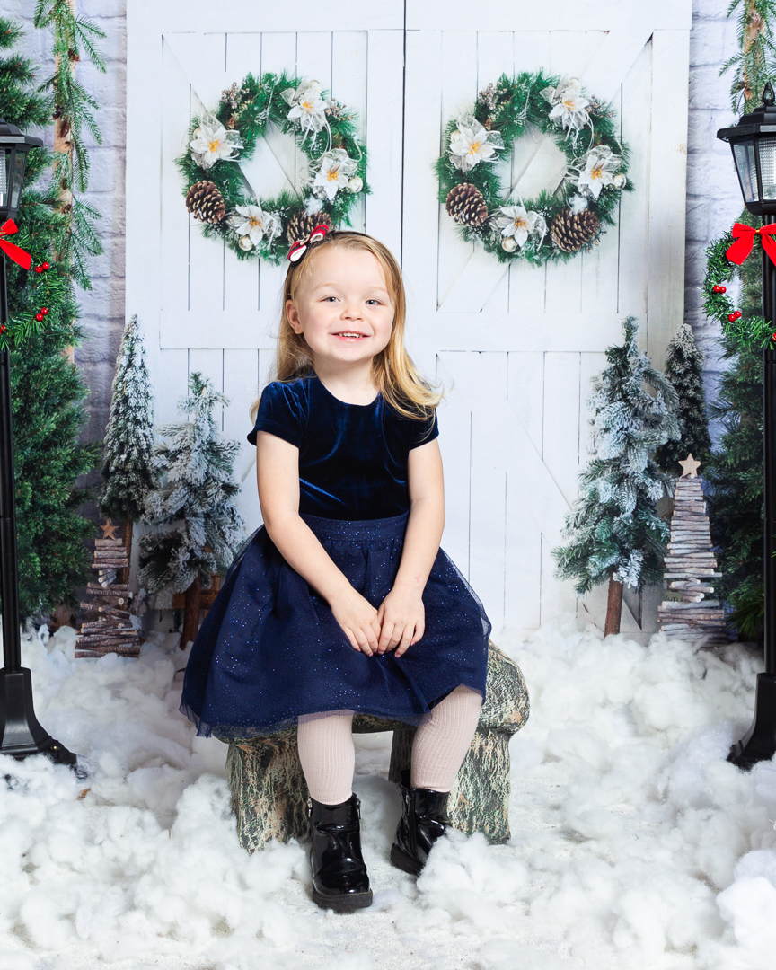 a little girl in a blue dress sits on a mini bench in a snow scene photo shoot