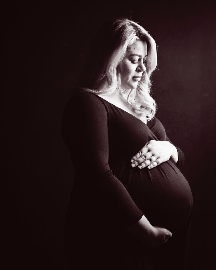 Expectant mother posing in a dark, moody studio setting, highlighting her baby bump.