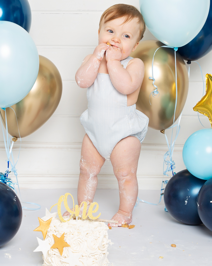 A little boy stands in front of some balloons with his birthday cake at a professional first birthday photo shoot