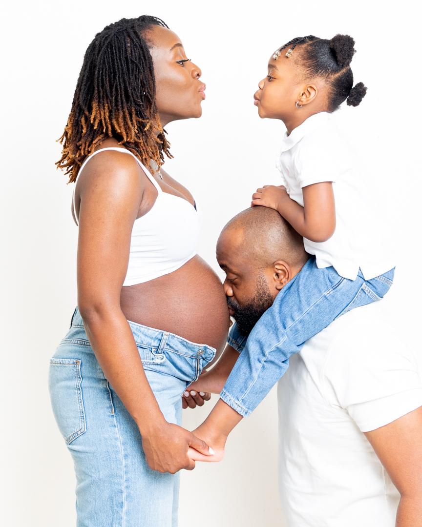 A little girl sits on her dad's shoulders and blows a kiss at her pregnant mum who is standing by them both