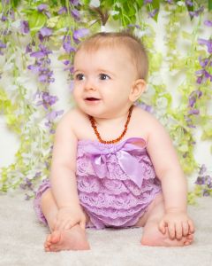 A baby is sitting in a lilac frilly outfit in front of a flower wall at a professional photoshoot at my West Sussex photography Studio