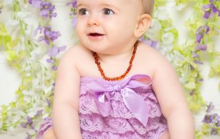 A baby poses in a lilac frilly outfit in a photographic studio in front of a mauve and white flower wall backdrop.