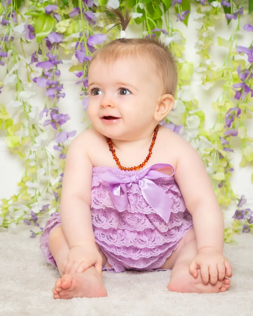 A baby poses in a lilac frilly outfit in a photographic studio in front of a mauve and white flower wall backdrop.