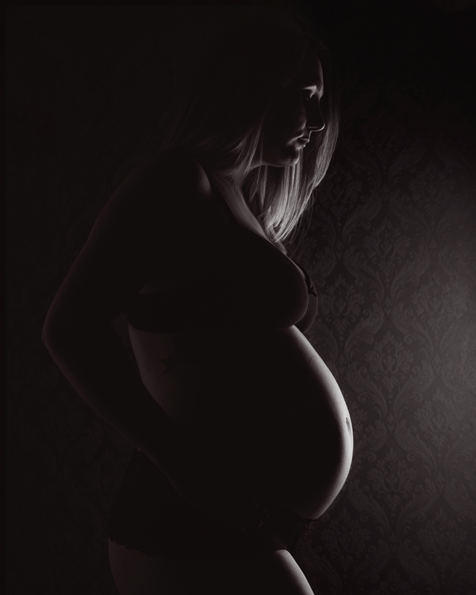 A dark black and white image of a pregnant women with the light just highlighting the edge of her face and bump