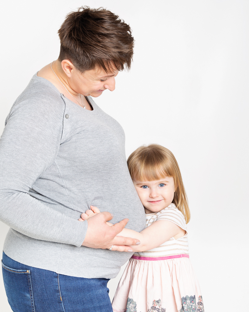 A little girl snuggles up to her mums pregnant belly in a colour studio image