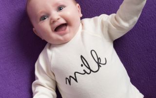 a smiling baby in a romper that says milk against a purple backdrop