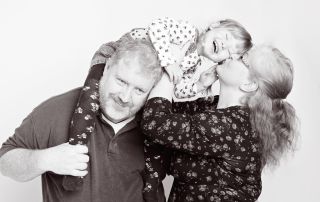 a black and white image of a little girl sitting on her dad's shoulders and leaning over for a kiss from her mum