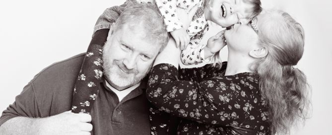 a black and white image of a little girl sitting on her dad's shoulders and leaning over for a kiss from her mum