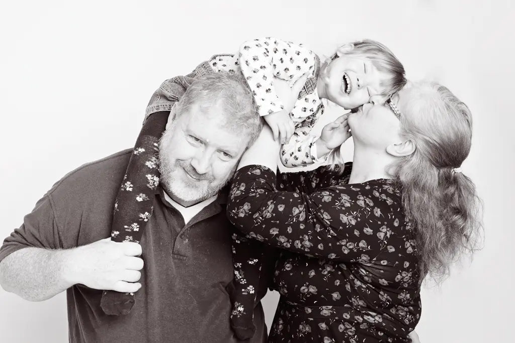 a black and white image of a little girl sitting on her dad's shoulders and leaning over for a kiss from her mum