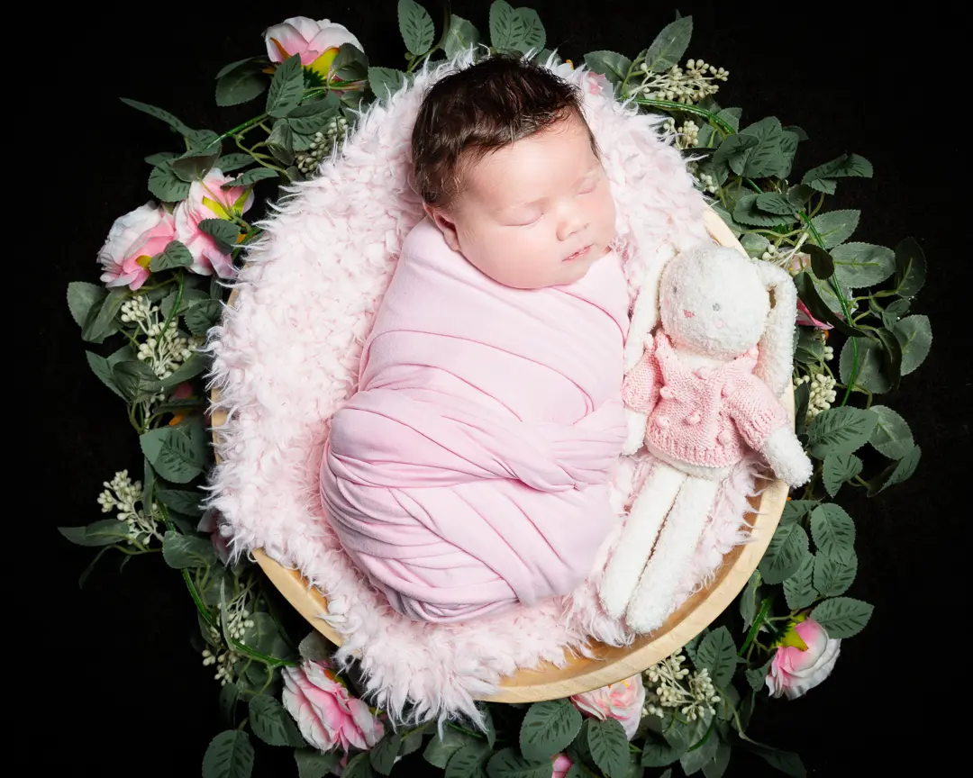 a newborn wrapped in pink sleeping in a bowl with a stuffed bunny toy