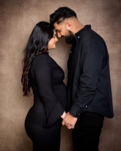 A couple, the woman is pregnant, pose facing each other with their heads touching and looking down at the bump