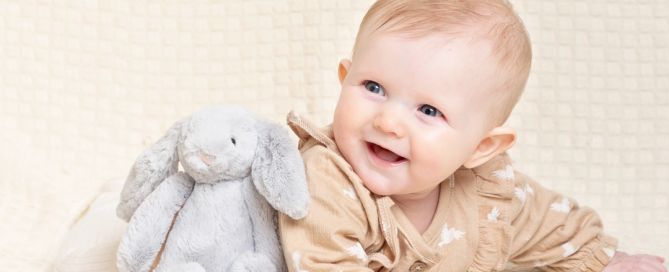 A Baby in a beige romper poses smiling with a toy bunny