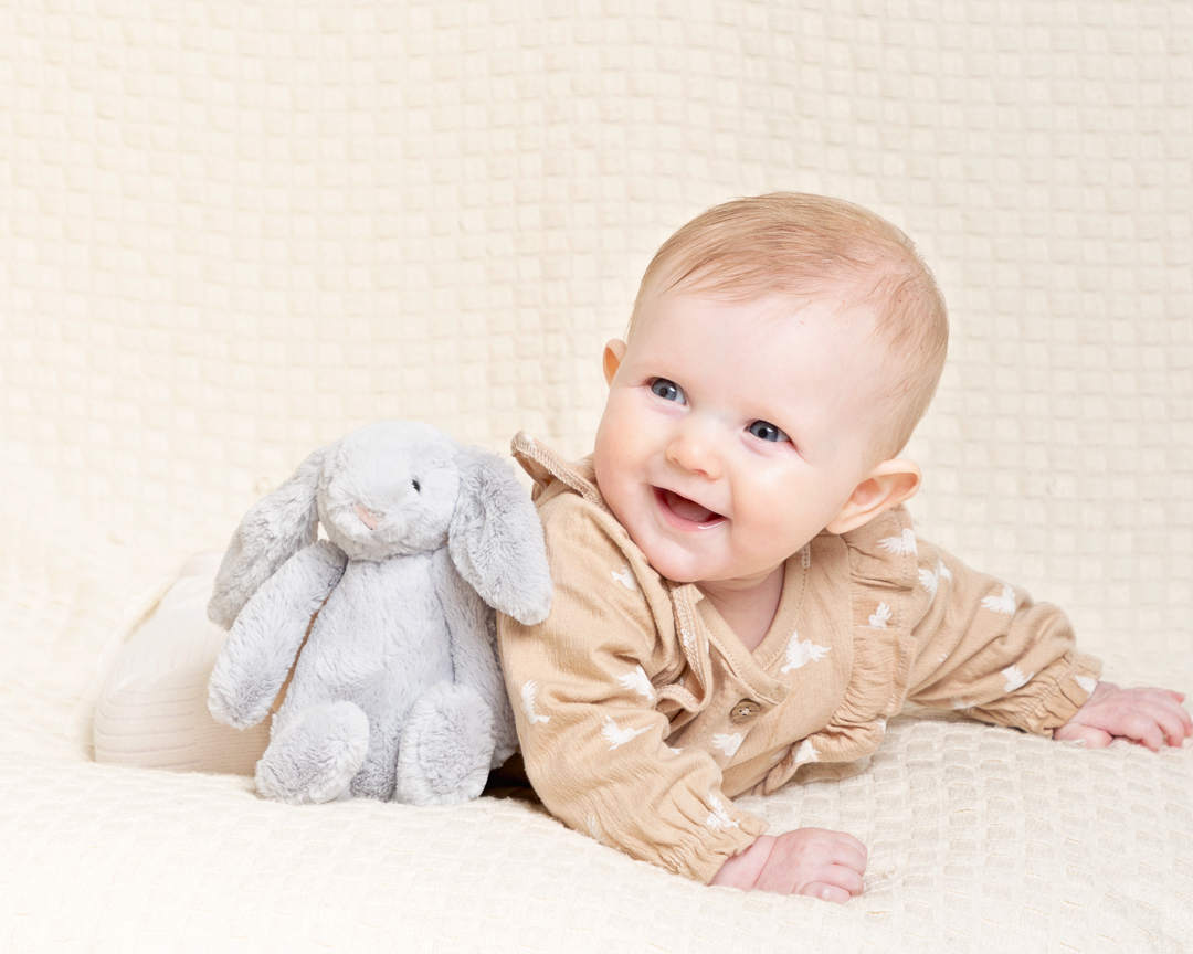 A Baby in a beige romper poses smiling with a toy bunny