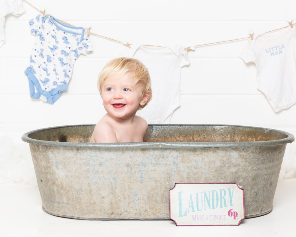 a little boy is smiling in a tin tub with a laundry sign and baby vests drying on a line