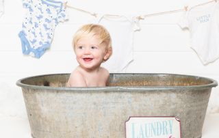 a little boy is smiling in a tin tub with a laundry sign and baby vests drying on a line