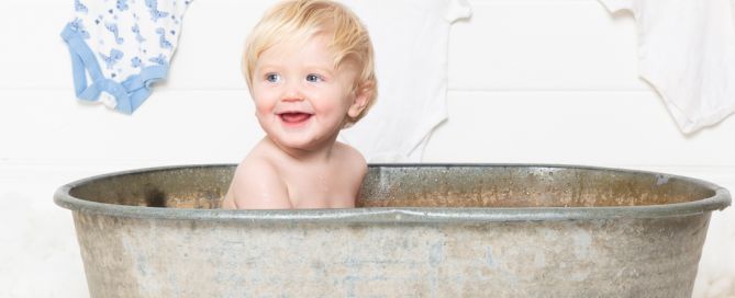 a little boy is smiling in a tin tub with a laundry sign and baby vests drying on a line