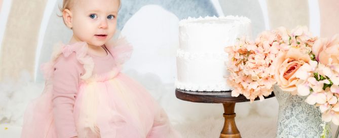 a baby in a pink dress poses with a cake in front of a pastel rainbow backdrop