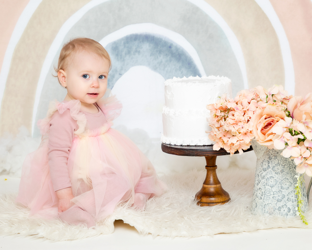 a baby in a pink dress poses with a cake in front of a pastel rainbow backdrop