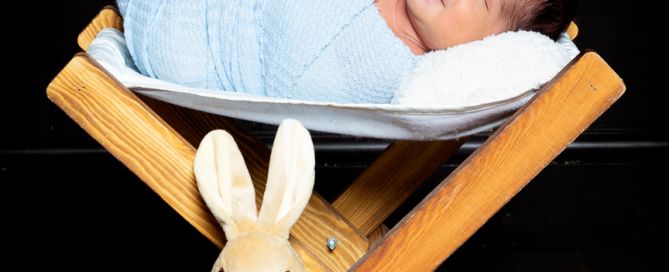 A sleeping newborn wrapped in blue is on a wooden hammock with a Peter Rabbit toy