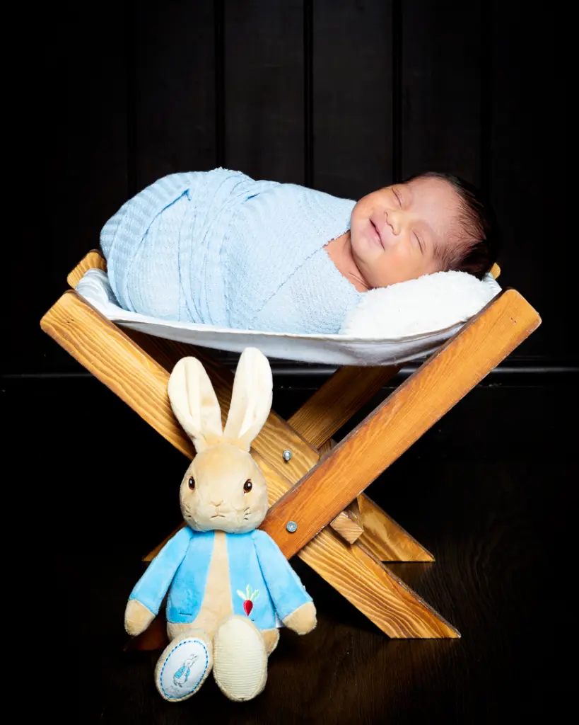 A sleeping newborn wrapped in blue is on a wooden hammock with a Peter Rabbit toy