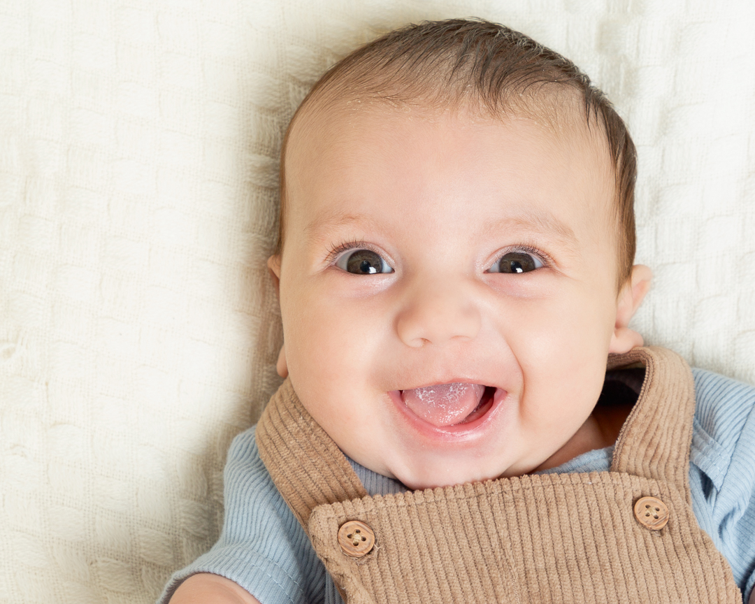 a happy baby boy in beige dungarees smiles up at the camera