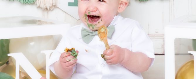 a little boy is really enjoying his cake at a first birthday photo shoot with a sage green theme