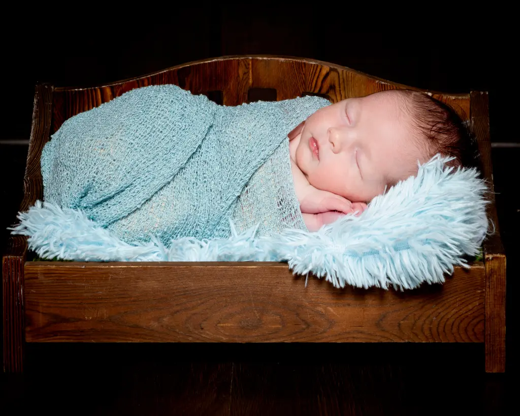 A newborn swaddled in pale blue sleeping on a miniature dark wood bed