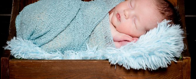 A newborn swaddled in pale blue sleeping on a miniature dark wood bed