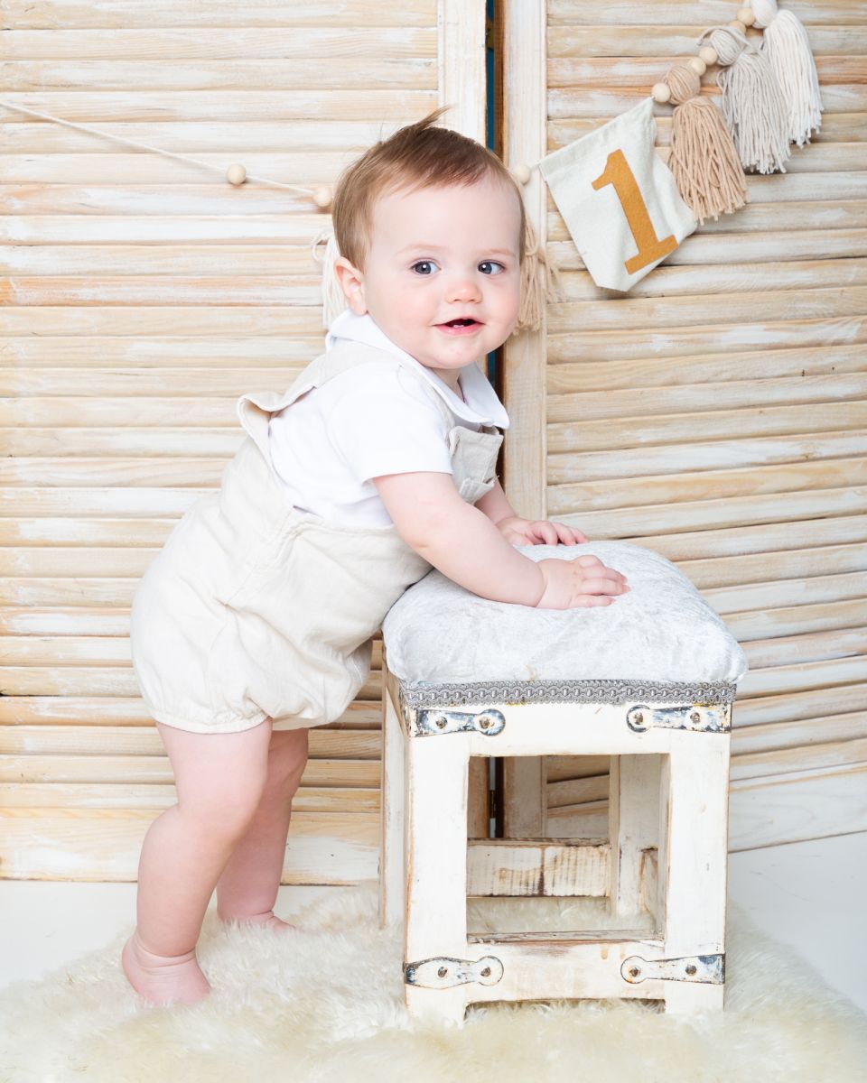 a little boy stands in a cream romper in front of a shutter with one bunting